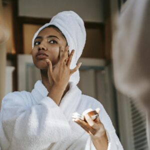 Crop young ethnic female in bathrobe with terry towel on head applying smooth cream on face while looking in mirror at home