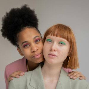Gentle black woman with pink eyeshadows embracing red haired female from behind and looking at camera against gray background