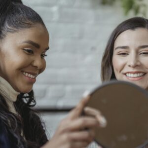 Two women smiling while holding a mirror indoors, showcasing joy and beauty.